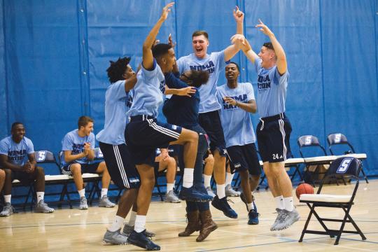 A group of athletes in blue NomaNation t-shirts and black shorts jump with excitement